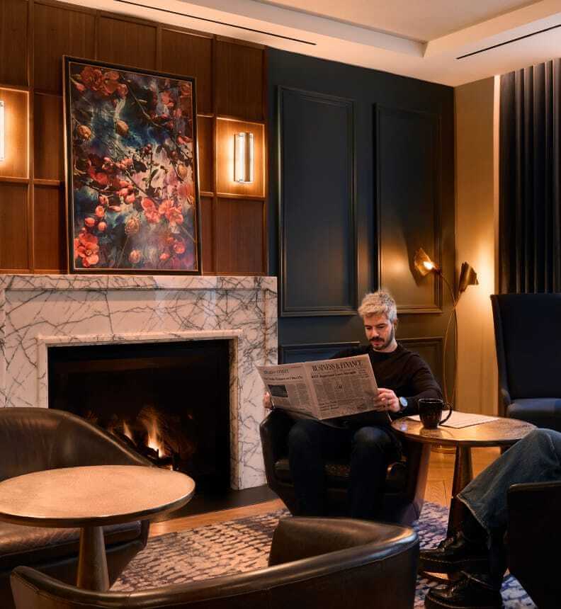 A man sits in a cozy lobby lounge with a fireplace, reading a newspaper.