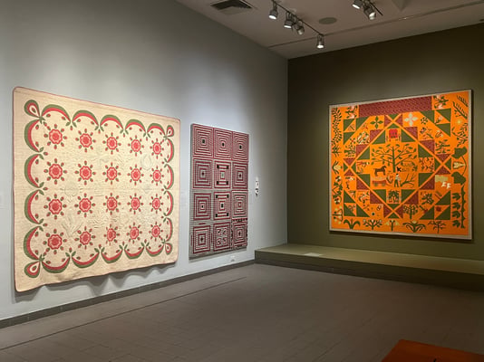 A spacious museum gallery displaying three large textile artworks mounted on gray walls.